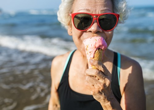 Senior woman wearing cool sunglasses and eating an ice cream cone on the beach.