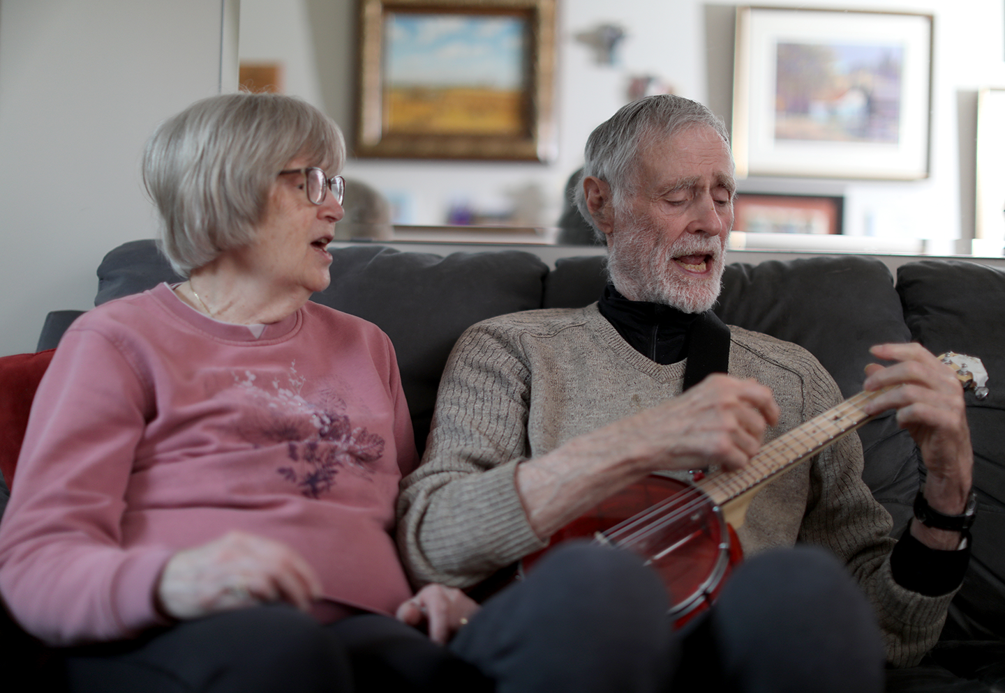 Margie and Brian sitting on a couch, while Brian plays the banjo.