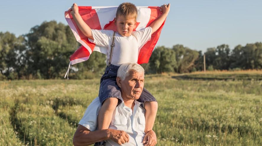 Young boy holding a Canadian flag and sitting on his grandfather's shoulders.