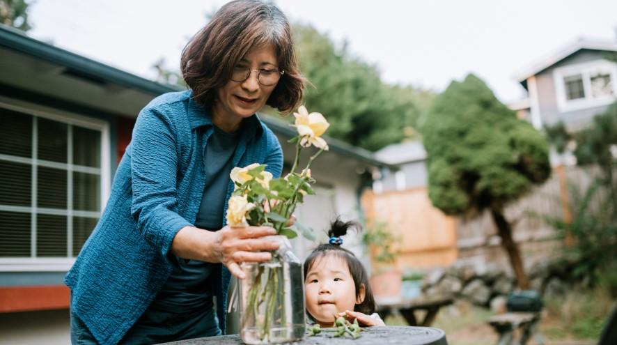 Senior woman putting flowers in a glass vase while her granddaughter watches.