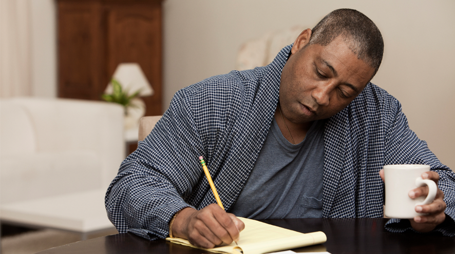 A man sits at a table holding a coffee cup and writing on a legal pad