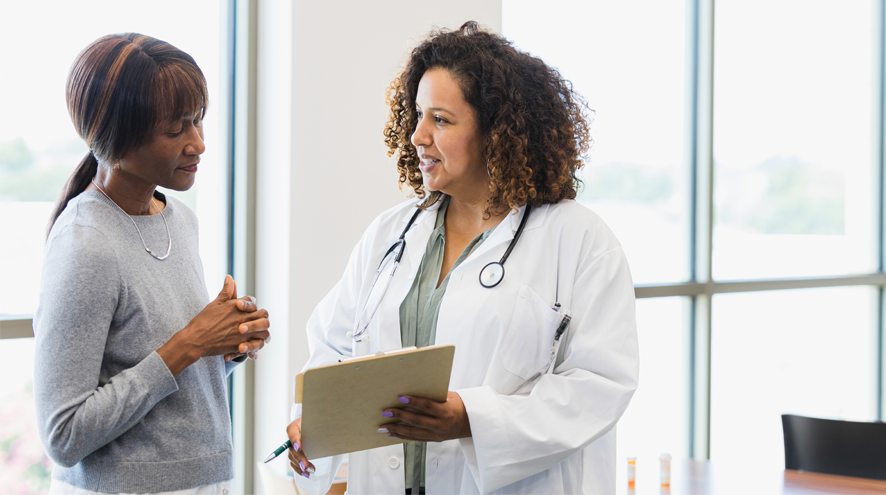 A woman wearing a labcoat and stethoscope discusses documents on a clipboard with a midlife woman