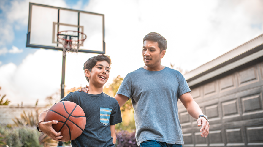 A boy holding a basketball walks with a man on a driveway basketball court