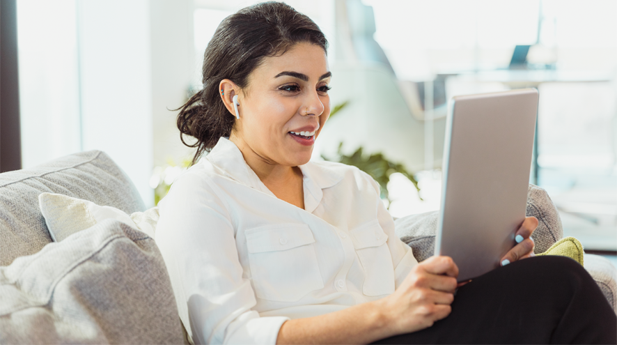 Midlife woman sits on a couch wearing earbuds and looking at a tablet