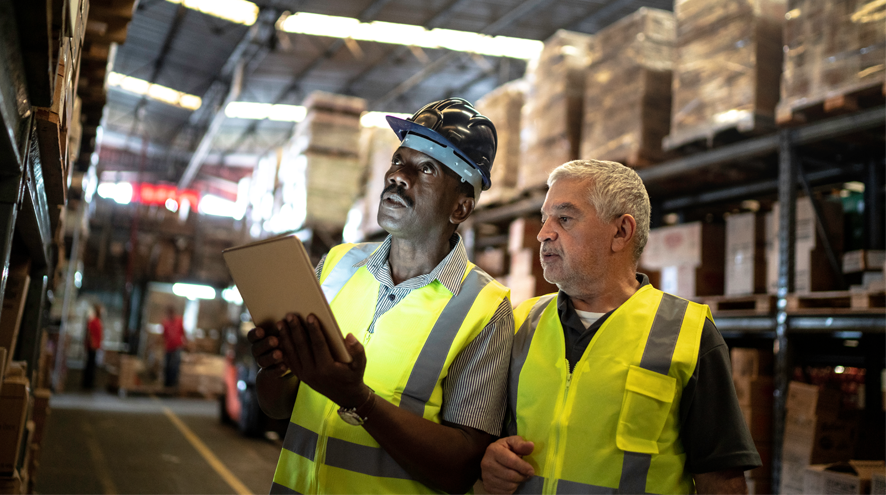 Two older midlife men wearing safety vests in a warehouse consult with a tablet computer