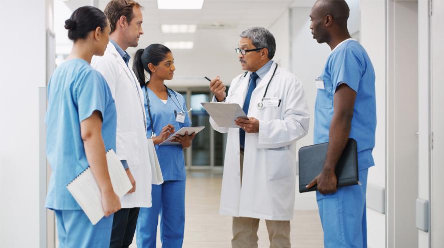 Five healthcare workers in scrubs and white jackets at a stand-up meeting
