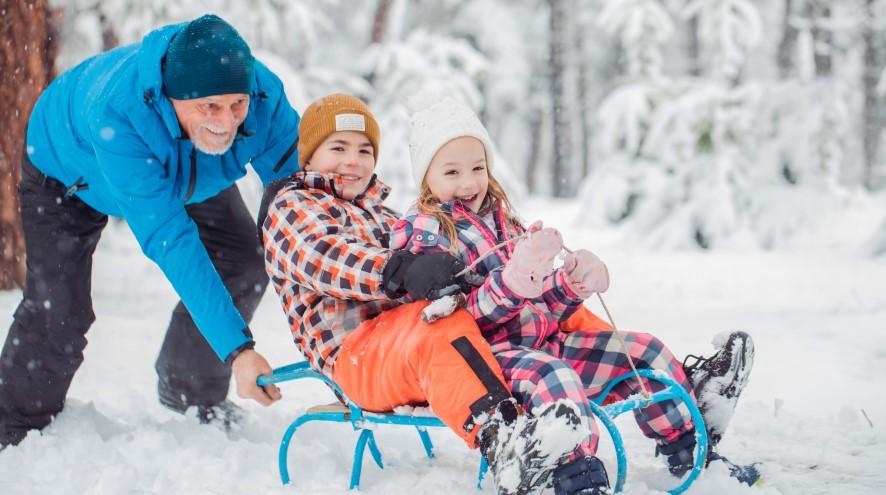 A happy grandfather is pushing his smiling children in their sleigh during a snowy winter scene.