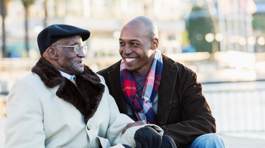 Older man in winter coat and cap sits next to midlife man in winter coat and scarf, they are smiling and talking
