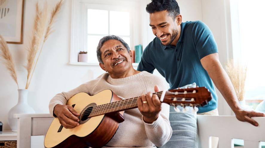 Older man playing guitar as younger man looks on, both smiling