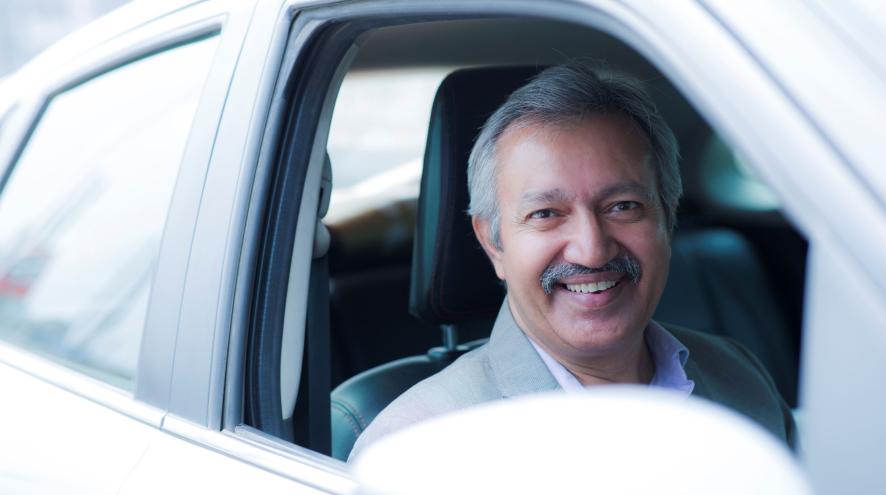 An older midlife man sits in the driver's seat of a white car, smiling