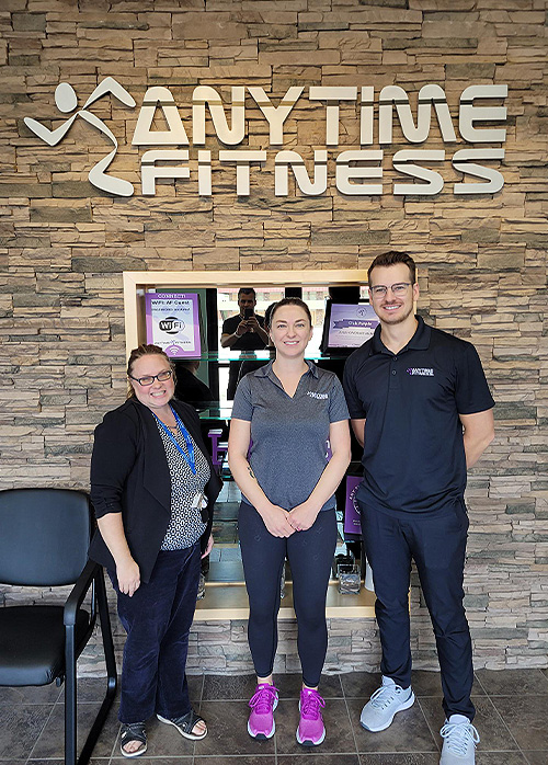 Our Dementia Community Coordinator with two Anytime Fitness staff members in front of the Anytime Fitness logo.