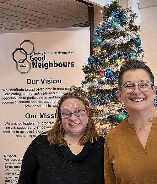 Our Dementia Community Coordinator and First Link Coordinator pictured in front of a Christmas tree and a Society for the Involvement of Good Neighbours banner.