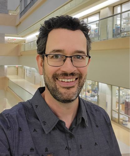 A headshot of Dr. John Howland. He has dark hair, and is wearing glasses and a dark grey shirt.