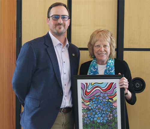 Two people standing next to each other smiling. The one on the left if wearing a navy suit jacket and glasses. The one on the right is wearing a black blazer, bright blue scarf, and is holding a framed Alzheimer Society award.