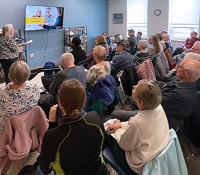 A person presenting to a crowd of people who are seated. The presenter is holding notes and motioning towards a screen that is displaying a presentation.