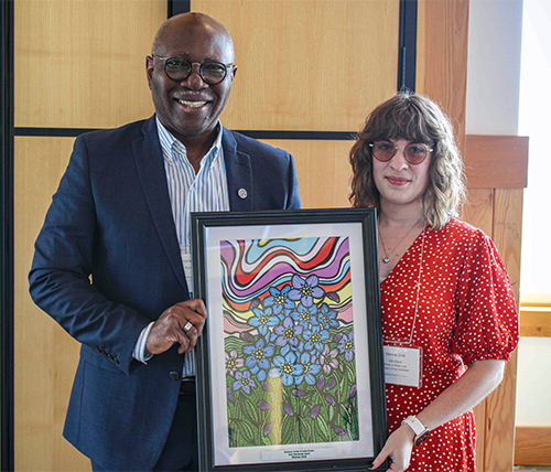 Two people standing next to each other smiling. The one on the left if wearing a navy suit jacket and the one on the right is wearing a red dress and holding a framed Alzheimer Society award.