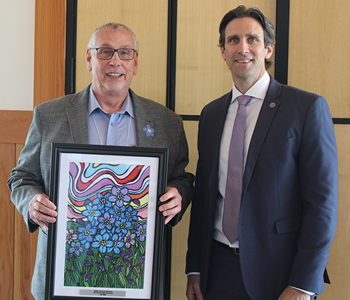 Two men standing next to each other smiling. The one on the left is holding a colourful Alzheimer Society of Saskatchewan framed award.
