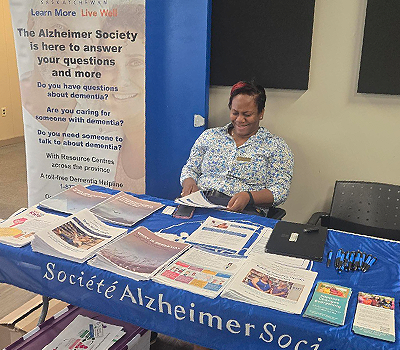 A smiling woman is seated at a display table that has many colourful booklets and handouts. The table covering says, "Alzheimer Society" and there is a banner in the background that says, "Alzheimer Society of Saskatchewan".
