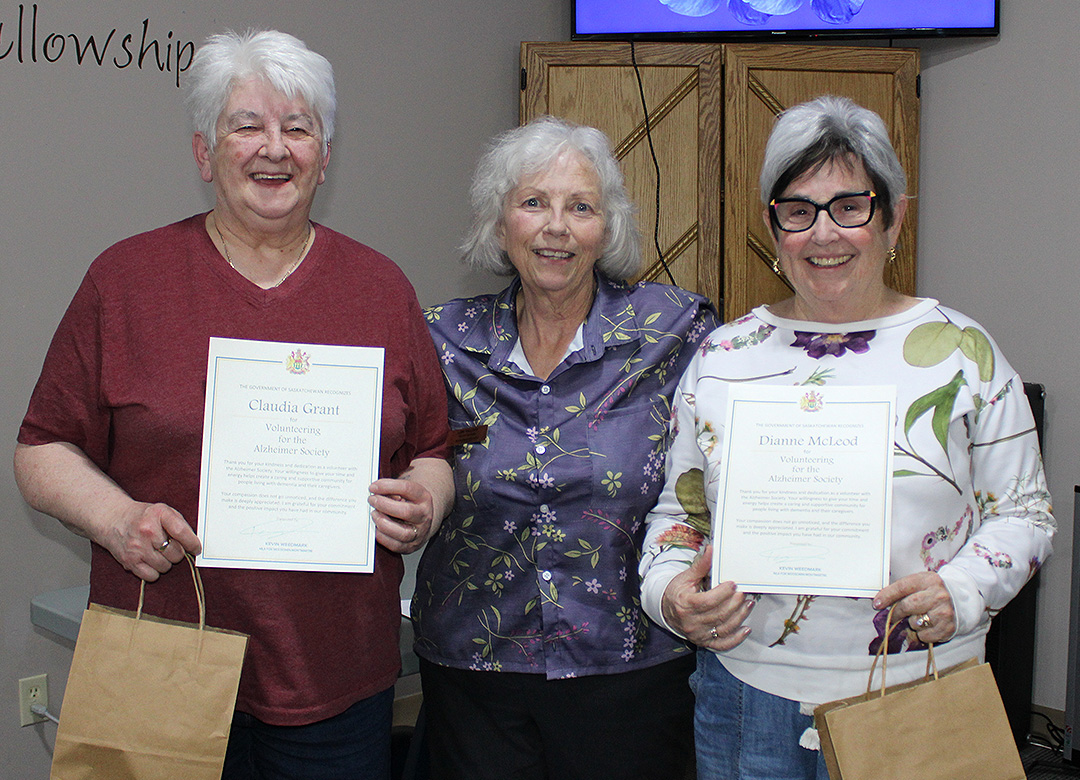 Three women standing in a line together and smiling. Two of the individuals are holding certificates and gift bags.