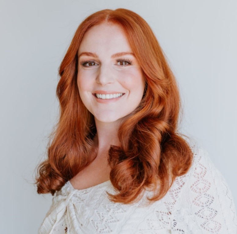 A woman with curled red hair is smiling at the camera. She is wearing a white knitted blouse.