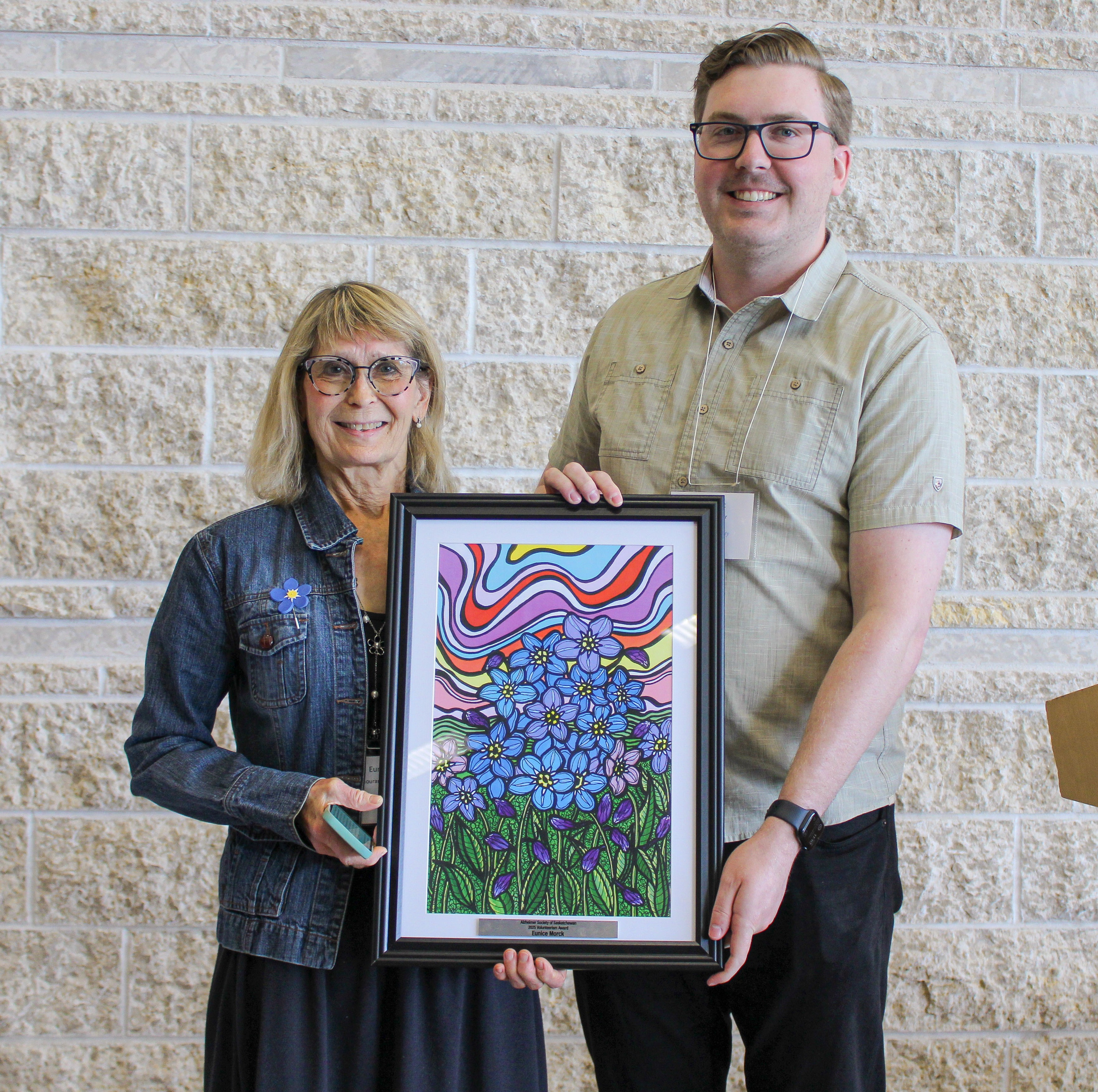 Two people standing indoors holding a framed artwork featuring blue flowers against a colorful abstract background.