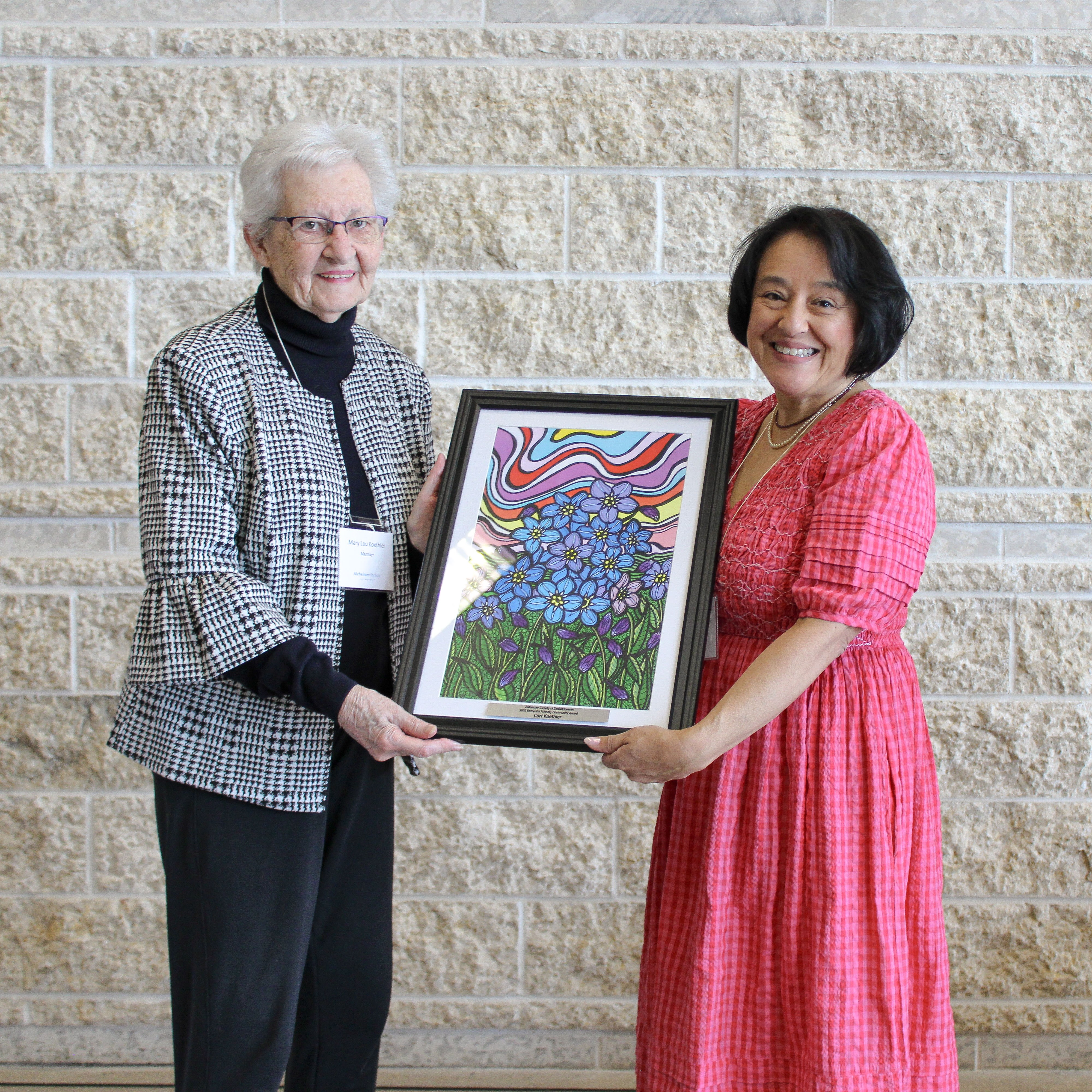 Two women standing indoors holding a framed artwork featuring blue flowers against a colorful abstract background.
