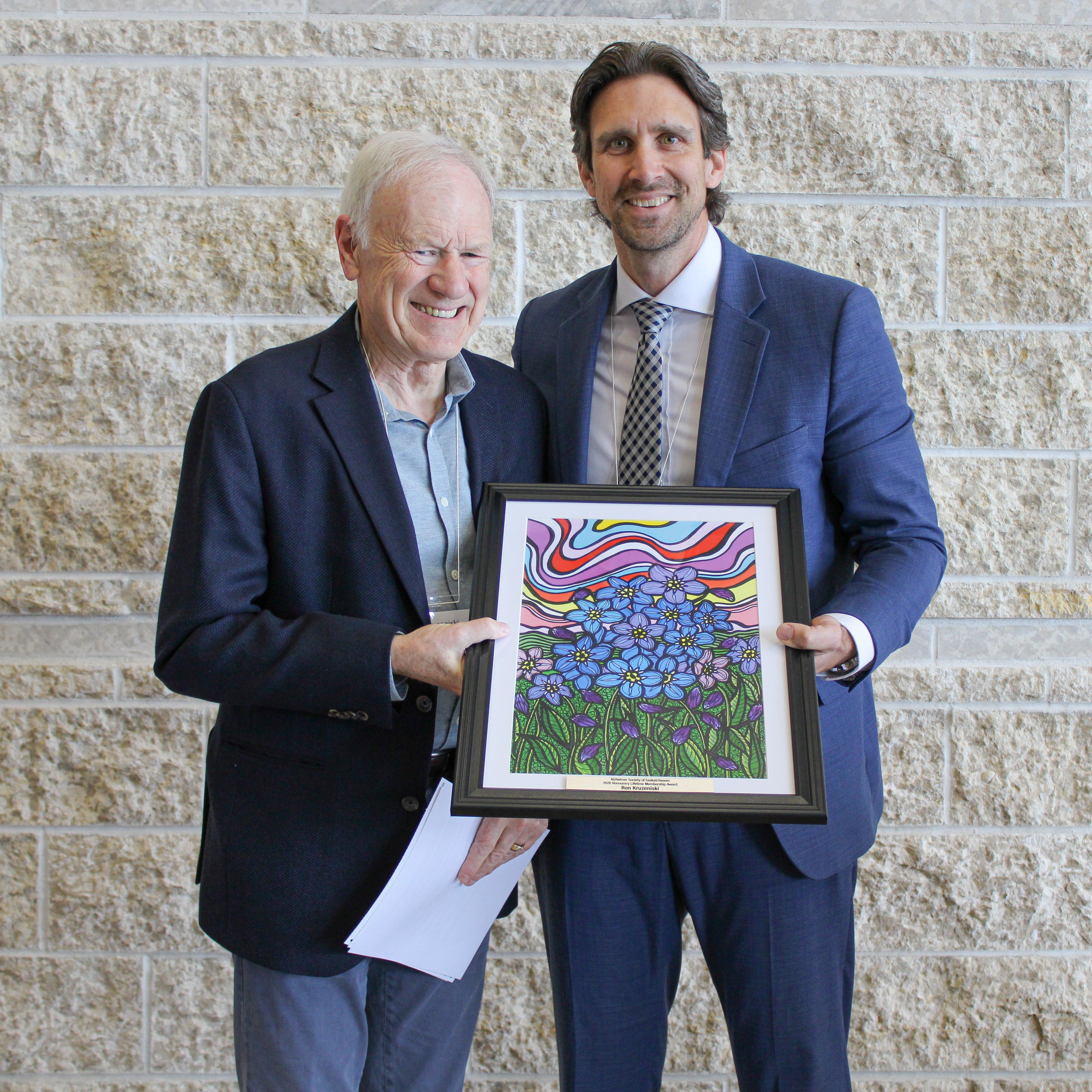 A man is presenting another man a colourful award plaque. They are both smiling.