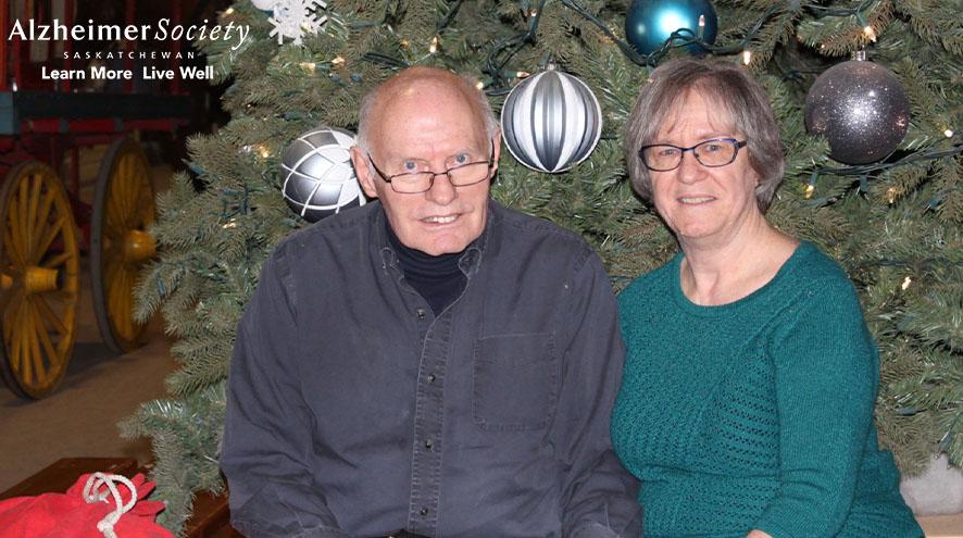 A husband and wife sitting together in front of a Christmas tree with large, round decorations.