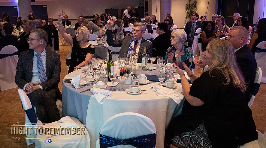 People in tuxedos and dresses sitting around a table with a white linen tablecloth. Two people are raising their hands to bid on an auction prize.