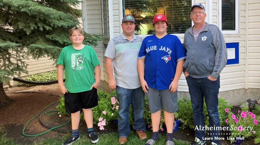 A father standing in front of a house with his three sons, who are all wearing different sports hats and shirts.