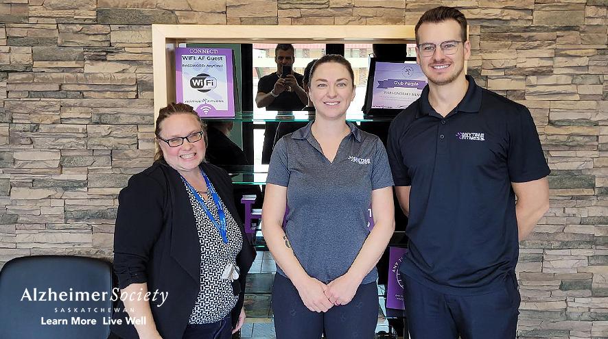 Our Dementia Community Coordinator standing next to two Anytime Fitness Yorkton representatives. Everyone is wearing black or dark grey colours and they are standing in front of a rock accent wall.