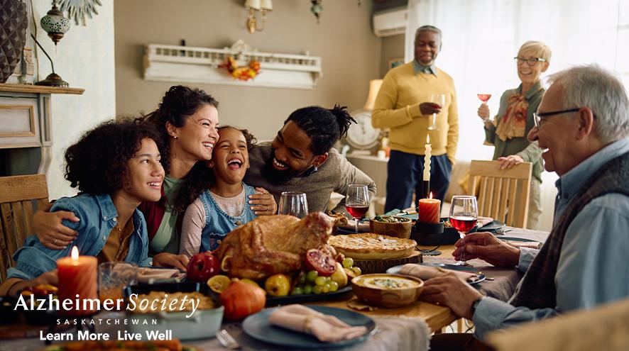 A mixed ages group of diverse people smiling and sitting around a kitchen table with a meal on it.