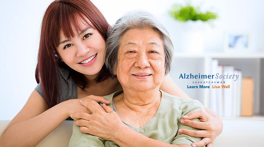 A younger woman has her hands lovingly wrapped around her aging mother, who is sitting down and smiling.