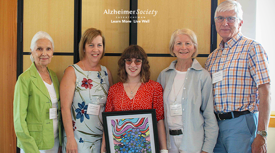 Four people standing around a person in a red dress and glasses. The person in the red dress is holding an Alzheimer Society of Saskatchewan framed award.