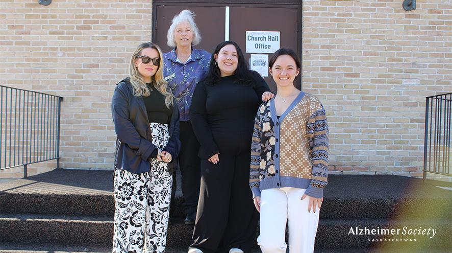 Four individuals standing outside of a church with a "Dementia in the Community Presentation" poster in the background.