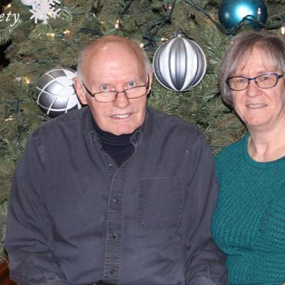A husband and wife sitting together in front of a Christmas tree with large, round decorations.