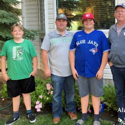 A father standing in front of a house with his three sons, who are all wearing different sports hats and shirts.
