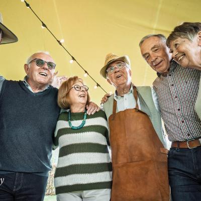 A group of people smiling with their arms around each other at on outdoor cookout. Some are wearing aprons.