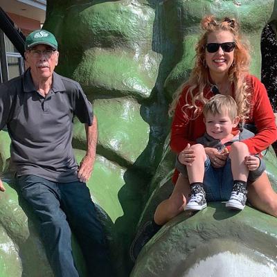 A man, his adult daughter, and his grandson are sitting on an oversized green dinosaur foot.