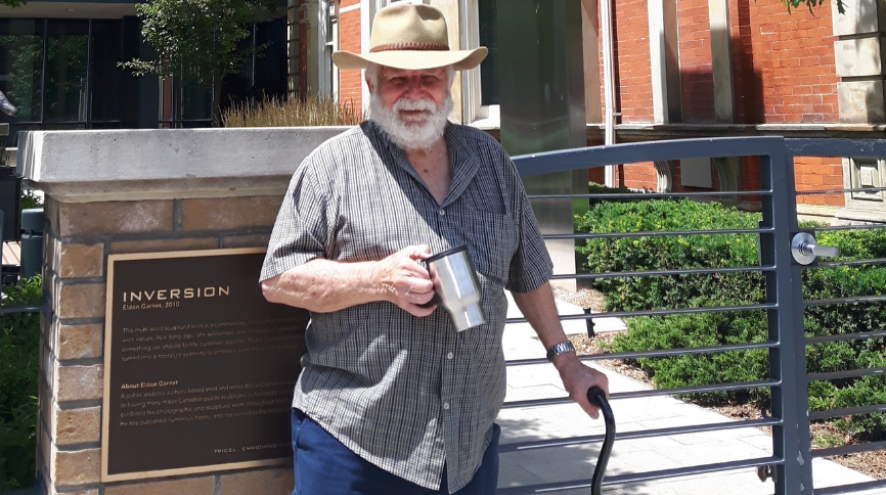 Picture of Bruce Howie in cowboy hat with coffee cup standing in front of a building holding onto a cane.