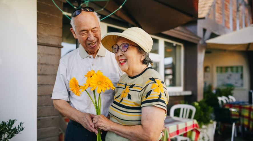 Older Asian couple with a woman wearing a hat, a yellow flowered sweater and holding yellow flowers