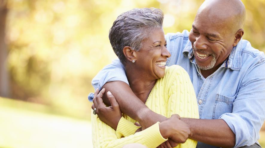 Black man with bald head wearing a blue shirt with his arms around a black woman with a yellow shirt. Both are laughing and smiling.