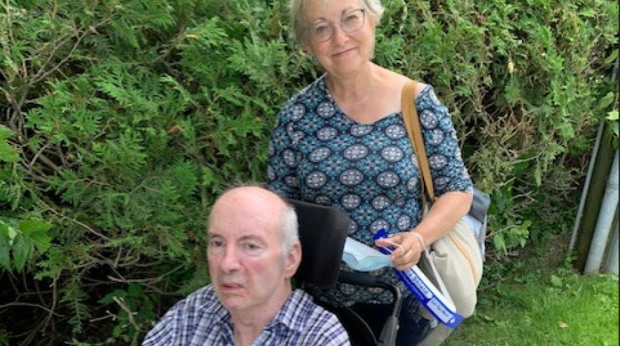 Older woman with short hair standing behind an older man in a wheelchair outside.