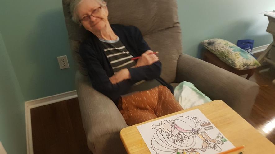 Older woman sitting in a brown chair with her arms crossed with a colouring picture in front of her on a table.