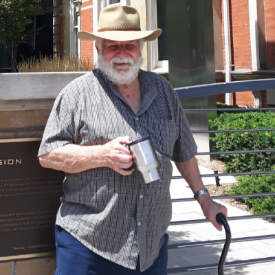 Picture of Bruce Howie in cowboy hat with coffee cup standing in front of a building holding onto a cane.