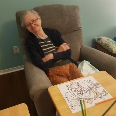 Older woman sitting in a brown chair with her arms crossed with a colouring picture in front of her on a table.