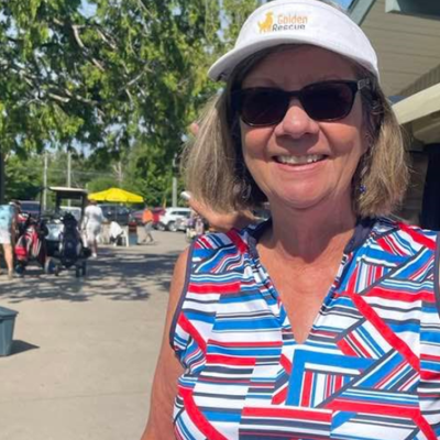 Head and shoulder picture of an older woman with shoulder length blond hair, wearing a golf visor and colourful shirt standing outside